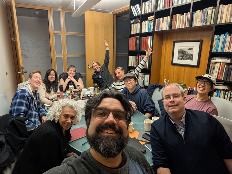 A group of enthusiastic, creative people gather around a table in a room lined with bookshelves. The person in the front takes a selfie of the whole group; several at the back of the photo are waving at the camera, arms outstretched; all are smiling.